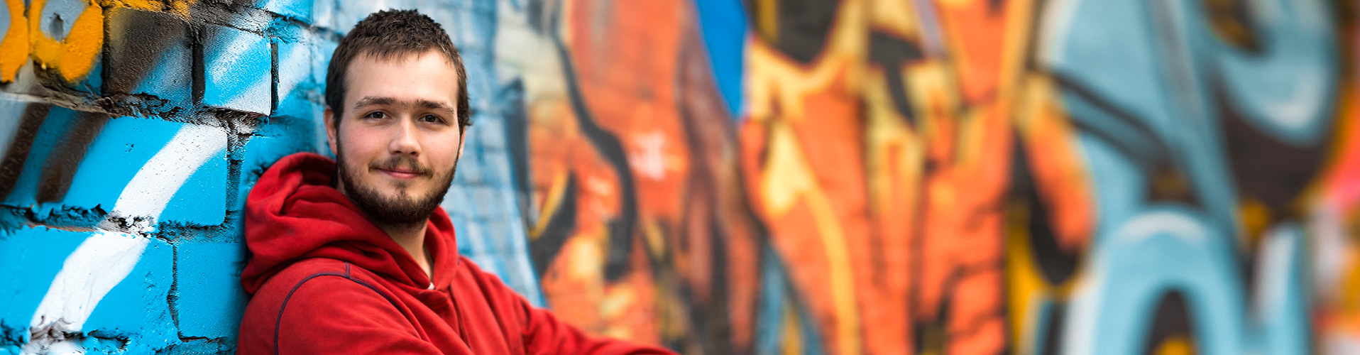 Junger Mann im roten Hoodie lehnt entspannt an einer bunt besprühten Graffiti-Wand und blickt in die Kamera.