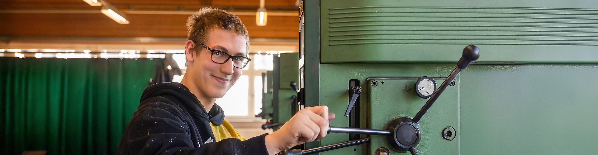 Jugendlicher mit Brille bedient eine große Metallmaschine in einer Werkstatt und schaut zur Kamera.