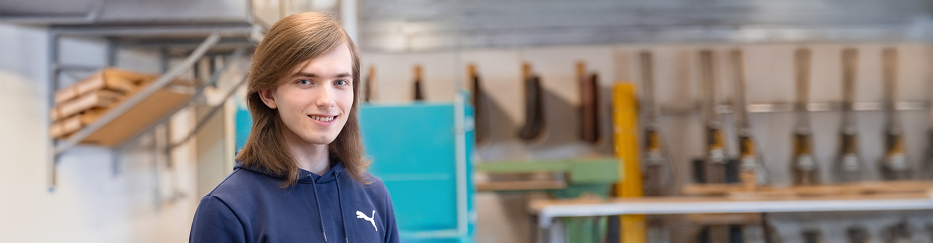 Jugendlicher mit langen Haaren steht in einer Werkstatt vor Maschinen und Werkzeugen und lächelt.
