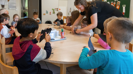 Kinder basteln gemeinsam an einem Tisch mit einer pädagogischen Fachkraft in einem Gruppenraum.
